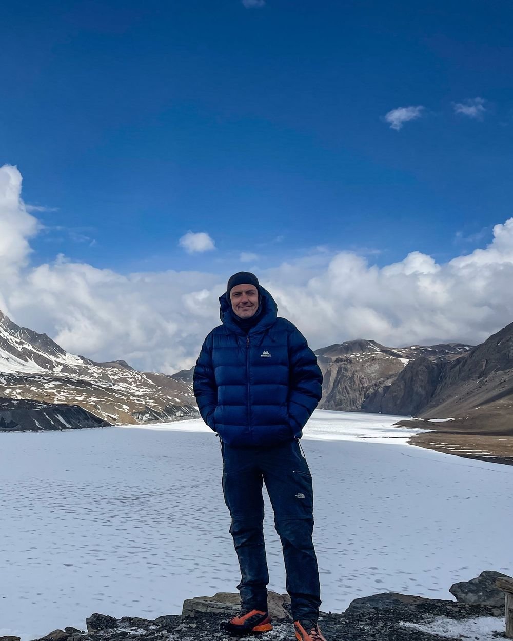 Alex Barratt in a blue down jacket, standing in front of a frozen lake and snow-covered mountains